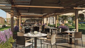 An outdoor dining area with round tables and chairs, a bar in the background, and a garden with purple flowers. - Borgo Pignano Florence (Florence)