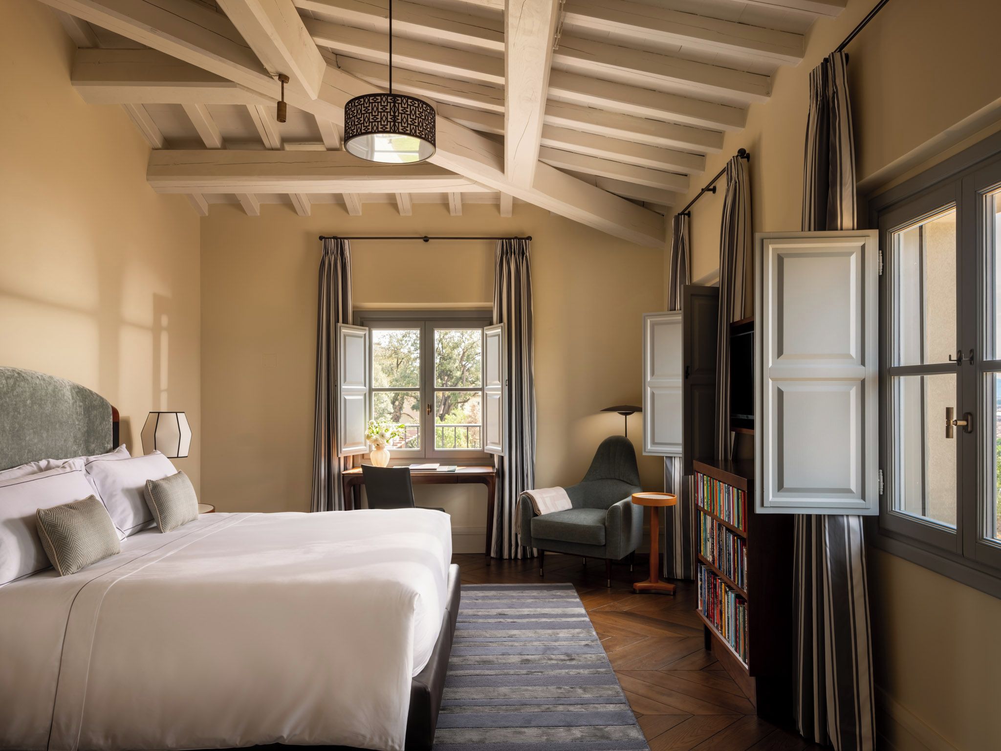A hotel room with a large bed, a desk, a chair, a bookshelf, and a window with curtains.