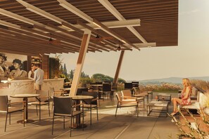 An outdoor seating area with tables and chairs, a bar with a bartender, and a view of the landscape.
