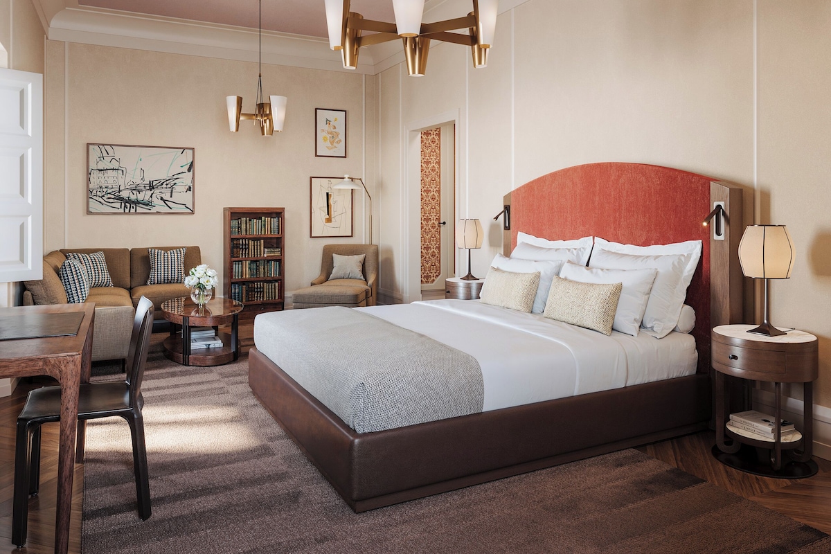 A hotel room with a large bed, a seating area with a sofa and armchair, a bookshelf, and a dining table.