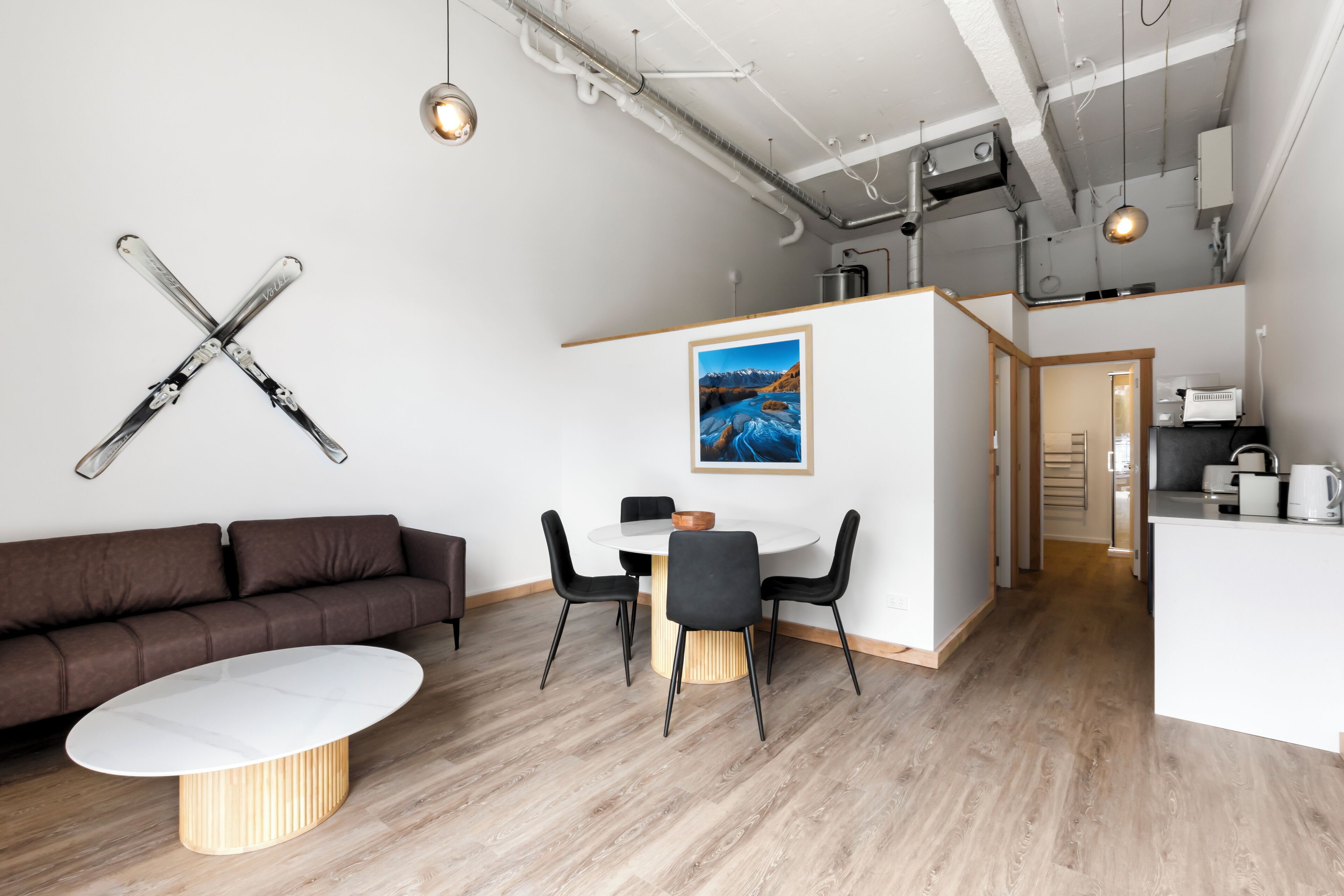 A modern living room with a brown sofa, a white coffee table, and a dining area with a white table and black chairs. There is a painting on the wall and a ski rack above the sofa.