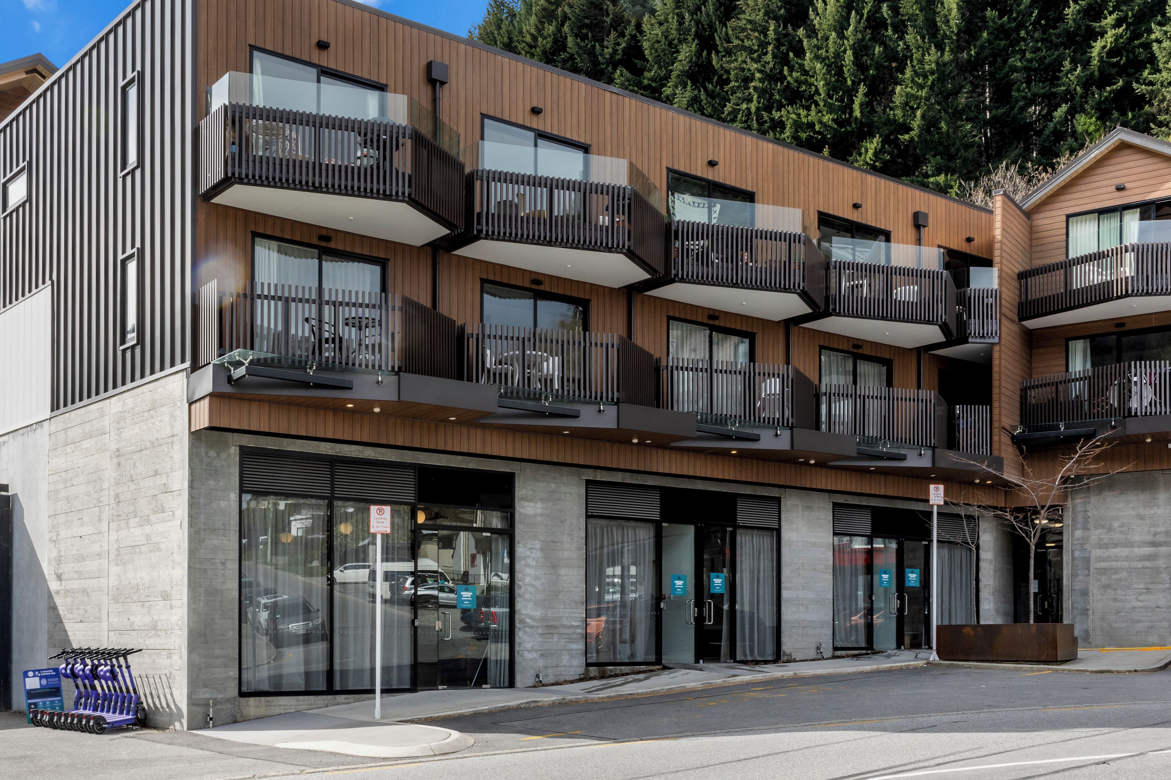Un edificio moderno de varios pisos con balcones, ventanas amplias y un estacionamiento con estación de carga para patinetes eléctricos azules.