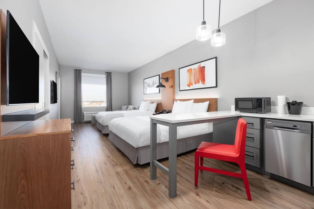 A modern hotel room with a large bed, a desk, a red chair, a TV, a microwave, and a dishwasher.