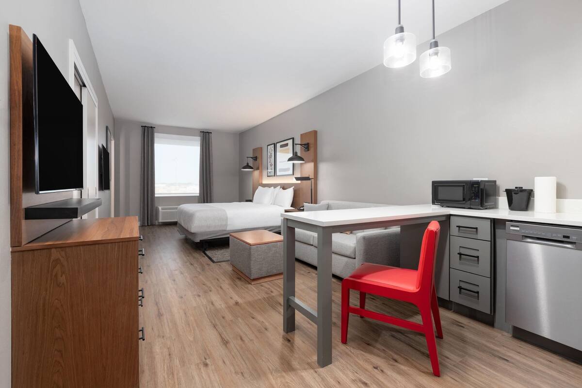 A modern hotel room with a bed, a desk, a red chair, a microwave, and a dishwasher.
