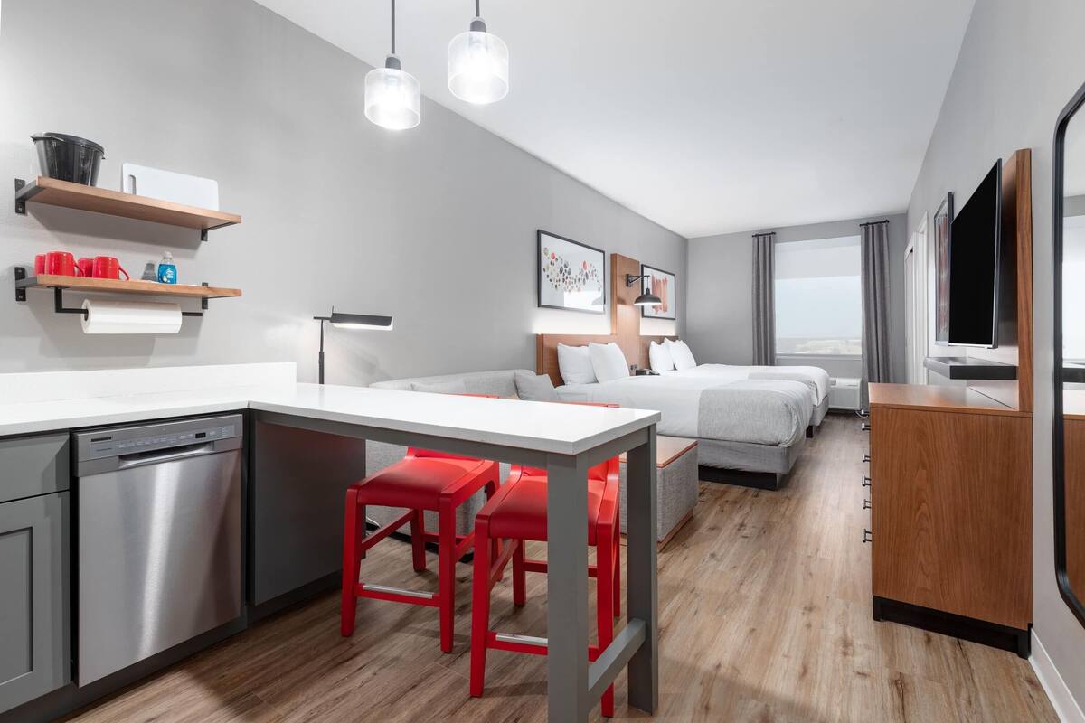 A modern kitchen with a dishwasher, a dining table with red chairs, and a grey sofa in the living area.
