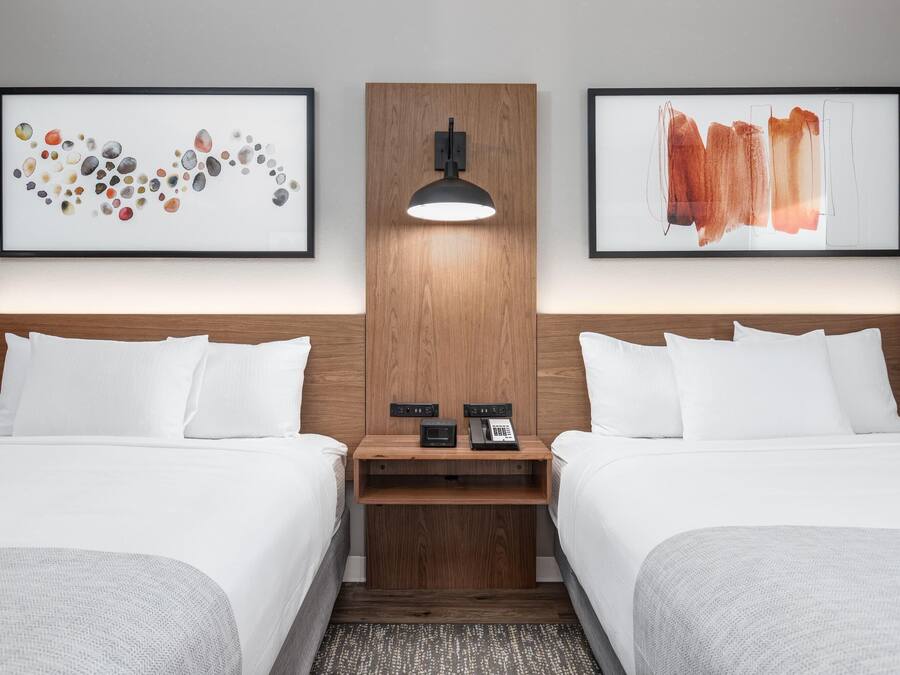 Ein Hotelzimmer mit zwei Betten, einem Holz-Kopfteil, einem Nachttisch mit Lampe sowie zwei gerahmten Kunstwerken an der Wand.