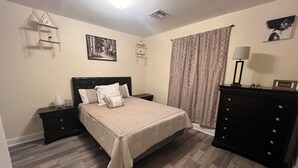 A bedroom with a bed, a dresser, a nightstand, a wall-mounted shelf, a framed picture, and a window with curtains. - Simplicity Stays NOLA (New Orleans)