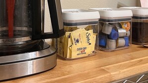A Krups coffee maker with a digital display, coffee pods, and mugs on a wooden shelf. - Simplicity Stays NOLA (New Orleans)
