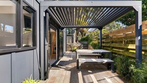 A covered patio area with a wooden table and benches, surrounded by a wooden fence and greenery.