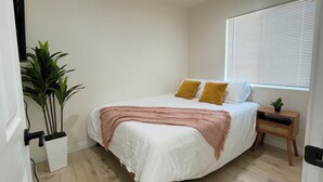 A bedroom with a bed, white walls, a window with blinds, a bedside table with a plant, and a wooden floor.