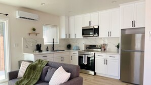 A modern kitchen with white cabinets, a stainless steel refrigerator, a microwave, and an oven. There is a gray sofa with a green throw blanket and white pillows in the living area.