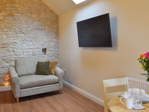 A cozy room with a stone wall, a grey sofa with cushions, a mounted TV, and a dining area with a table set for two.