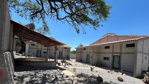 Pátio com dois edifícios, área de estar coberta e chão de cascalho com cactos.
