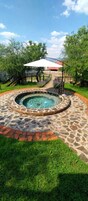 Una piscina de piedra circular con un borde de ladrillo, una zona de estar techada y un primer plano con césped.