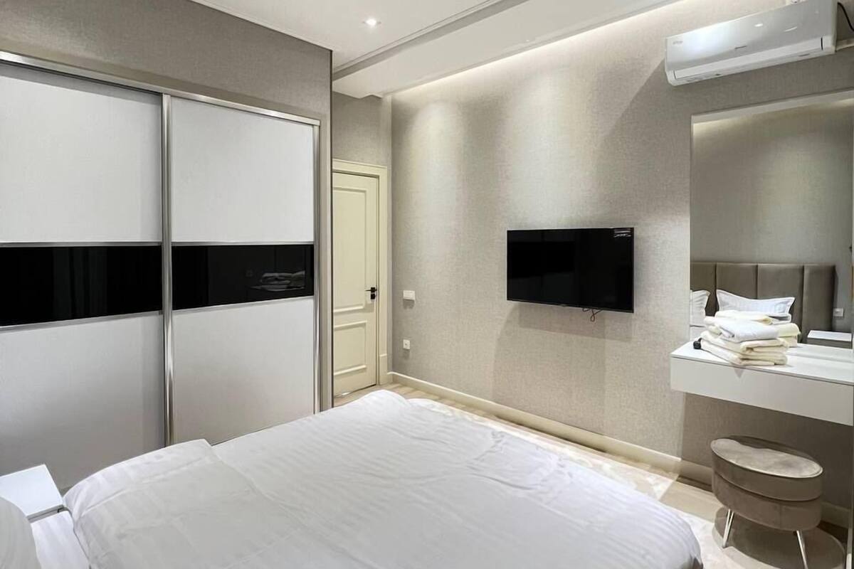 A modern bedroom with a large bed, a flat-screen TV mounted on the wall, a vanity with a mirror, and a sliding door closet.