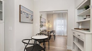 A modern kitchen with white cabinetry, a dining area with round tables and black chairs, and a large framed picture on the wall. - Iris Suite I Walk to Shops, Dining + Bay (Coronado)