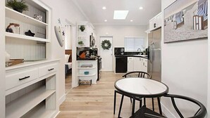 A modern kitchen with white cabinetry, a dining area with a round table and chairs, and a view of the living room in the background. - Iris Suite I Walk to Shops, Dining + Bay (Coronado)