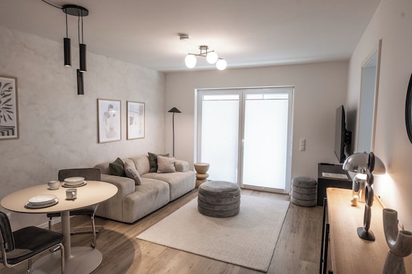 Uma sala de estar moderna com uma mesa de jantar redonda, um sofá e uma mesa de centro. Há quadros nas paredes, um abajur e uma TV fixada na parede.