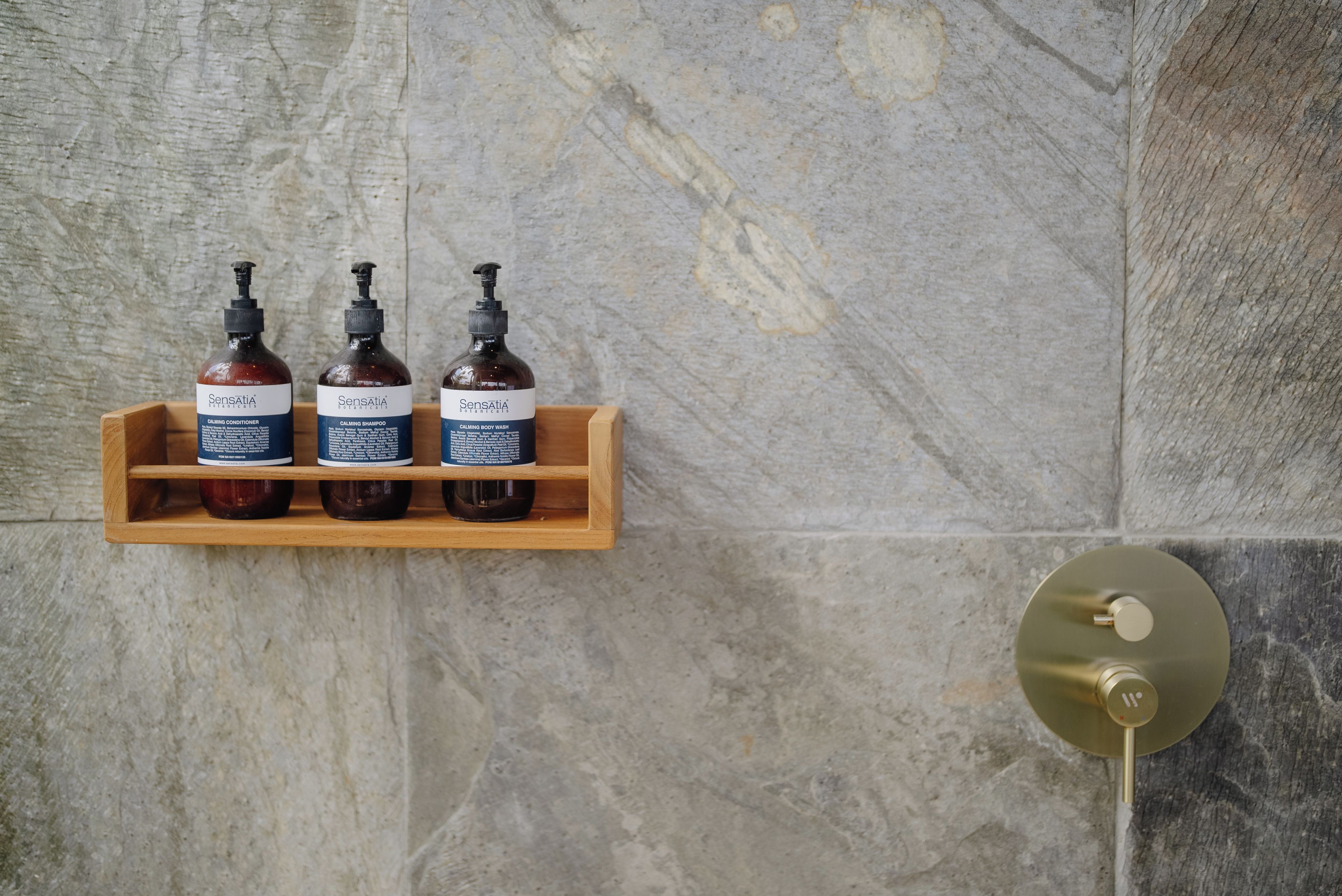 Une étagère en bois sur laquelle sont posées trois bouteilles de produits de la marque Sensatia Botanicals, le tout adossé à un mur en pierre.