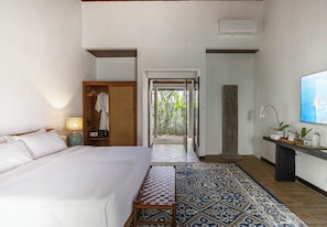 A hotel room with a bed, bedside table, lamp, wardrobe, and a view of a garden through an open door.