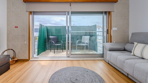 Una sala moderna con un sofá gris, un tapete redondo y un ventanal corredero que da a un área de estar exterior con dos sillas.