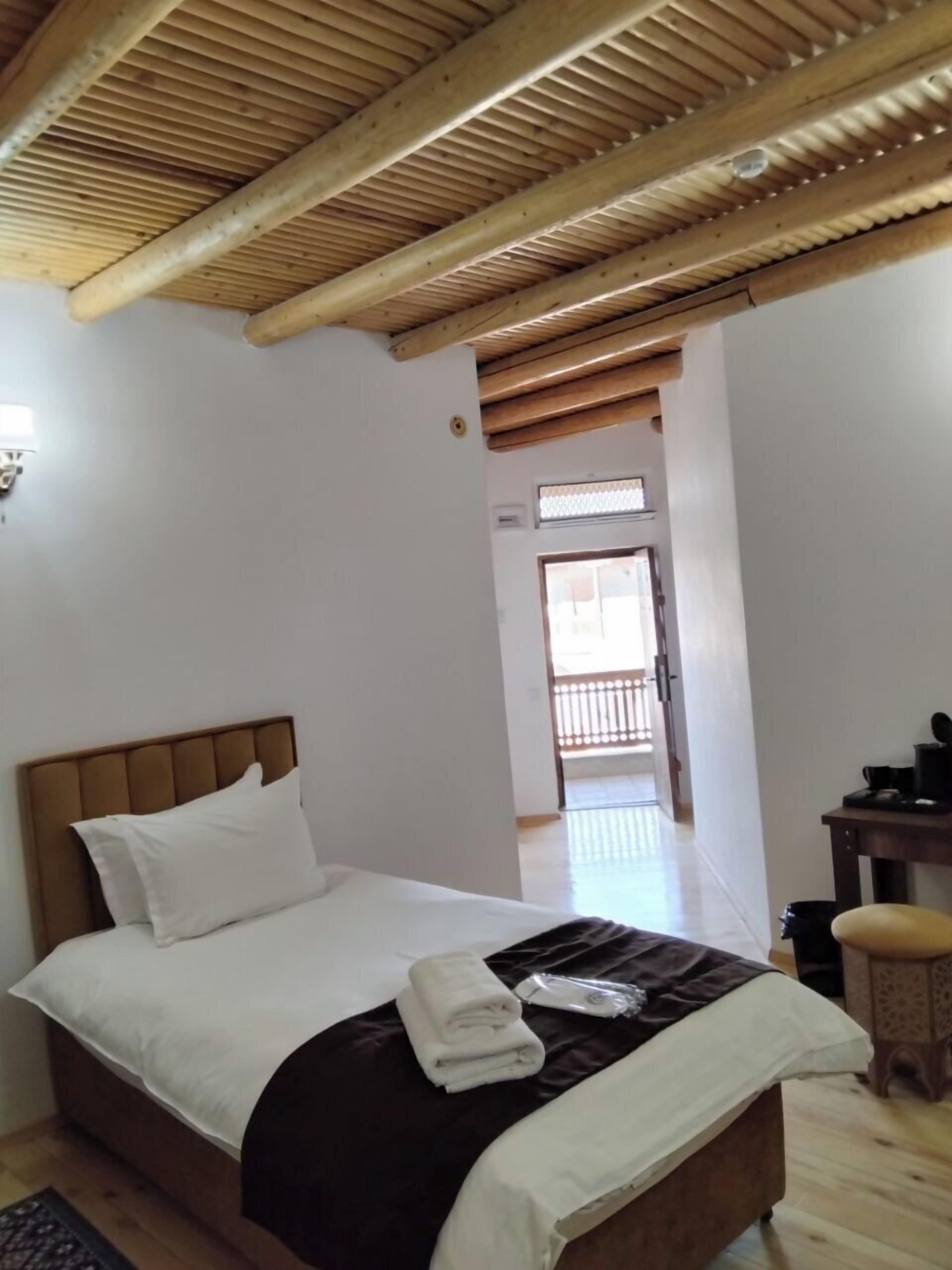 A bedroom with a wooden ceiling, a bed with white and brown bedding, a small table with a chair, and a view of a balcony.