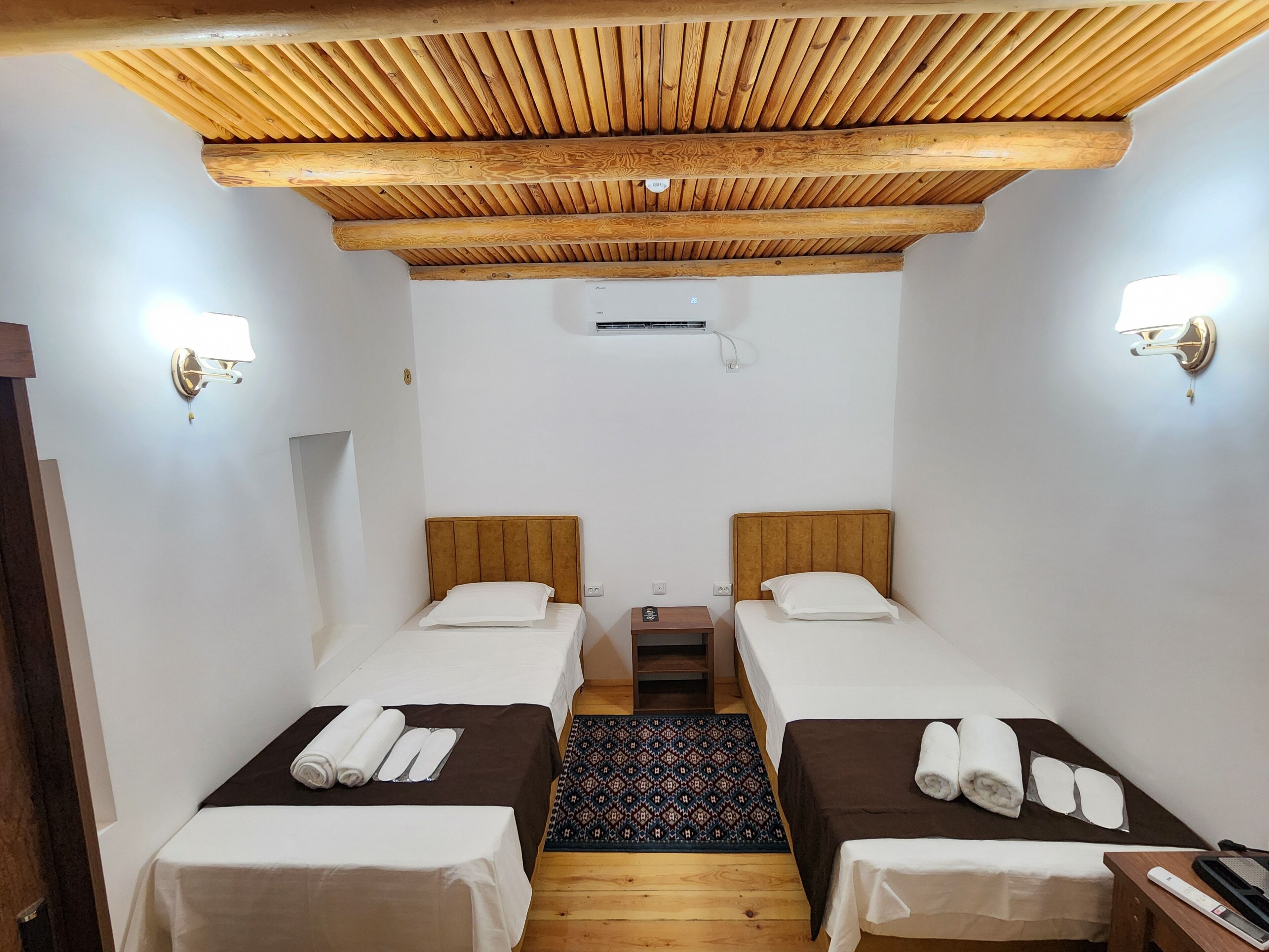 A room with two beds, wooden ceiling beams, wall-mounted lights, and a small table with a rug.