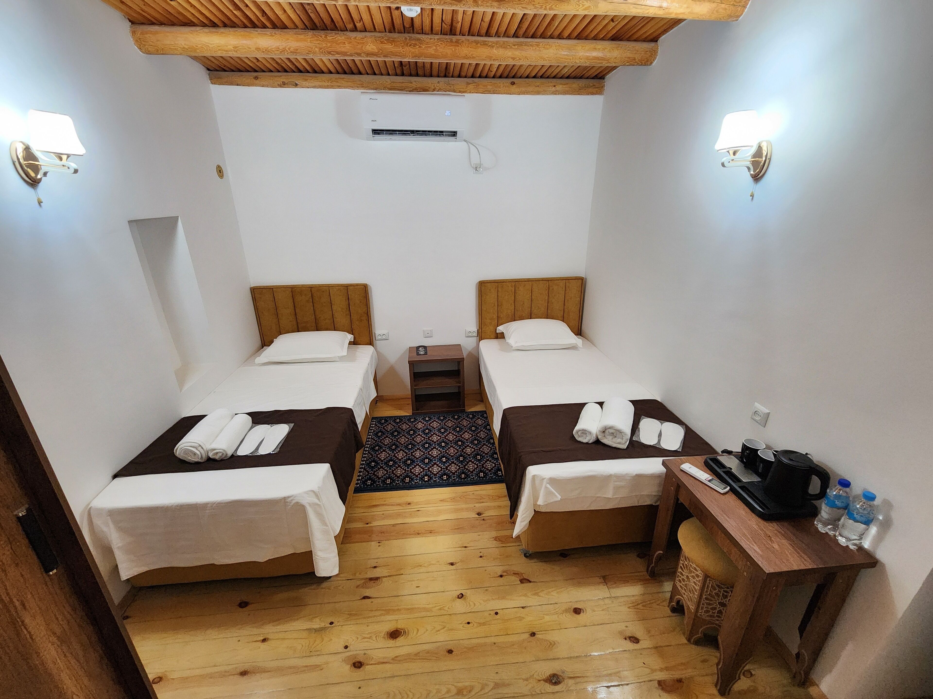 A small, cozy room with two single beds, a wooden ceiling, and a small table with a kettle and water bottles.