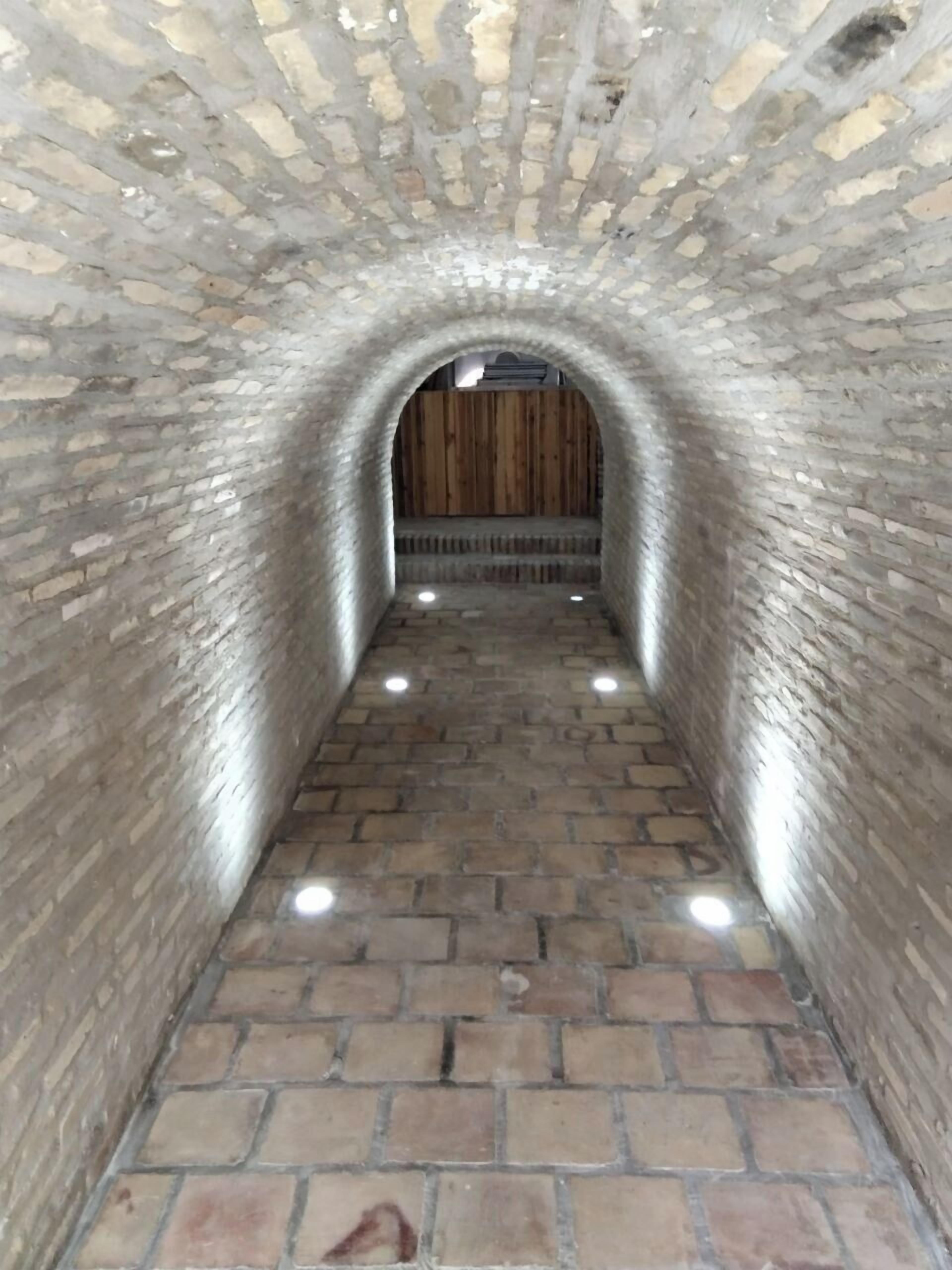 A stone-walled corridor with a wooden door at the end, leading to a room with a tiled floor and a bench.