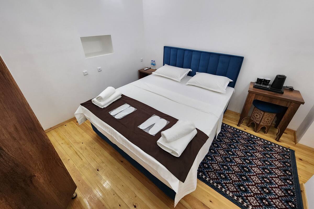 A neatly made bed with white linens and a brown accent, a bedside table with a lamp, and a patterned rug on the wooden floor.