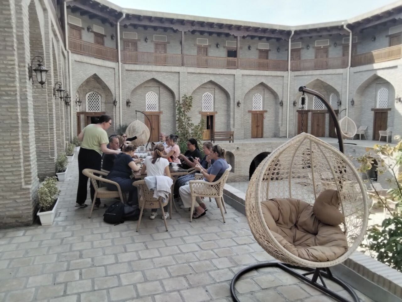 Une cour où un groupe de personnes est assis à une table pour dîner, entourée de murs en briques percés d’arches et de fenêtres et de portes en arc de cercle.