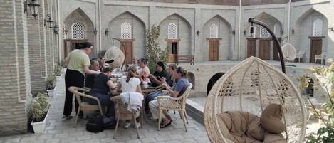 Un patio con un grupo de personas cenando en una mesa, rodeado de muros de ladrillo con ventanas y puertas de arco.