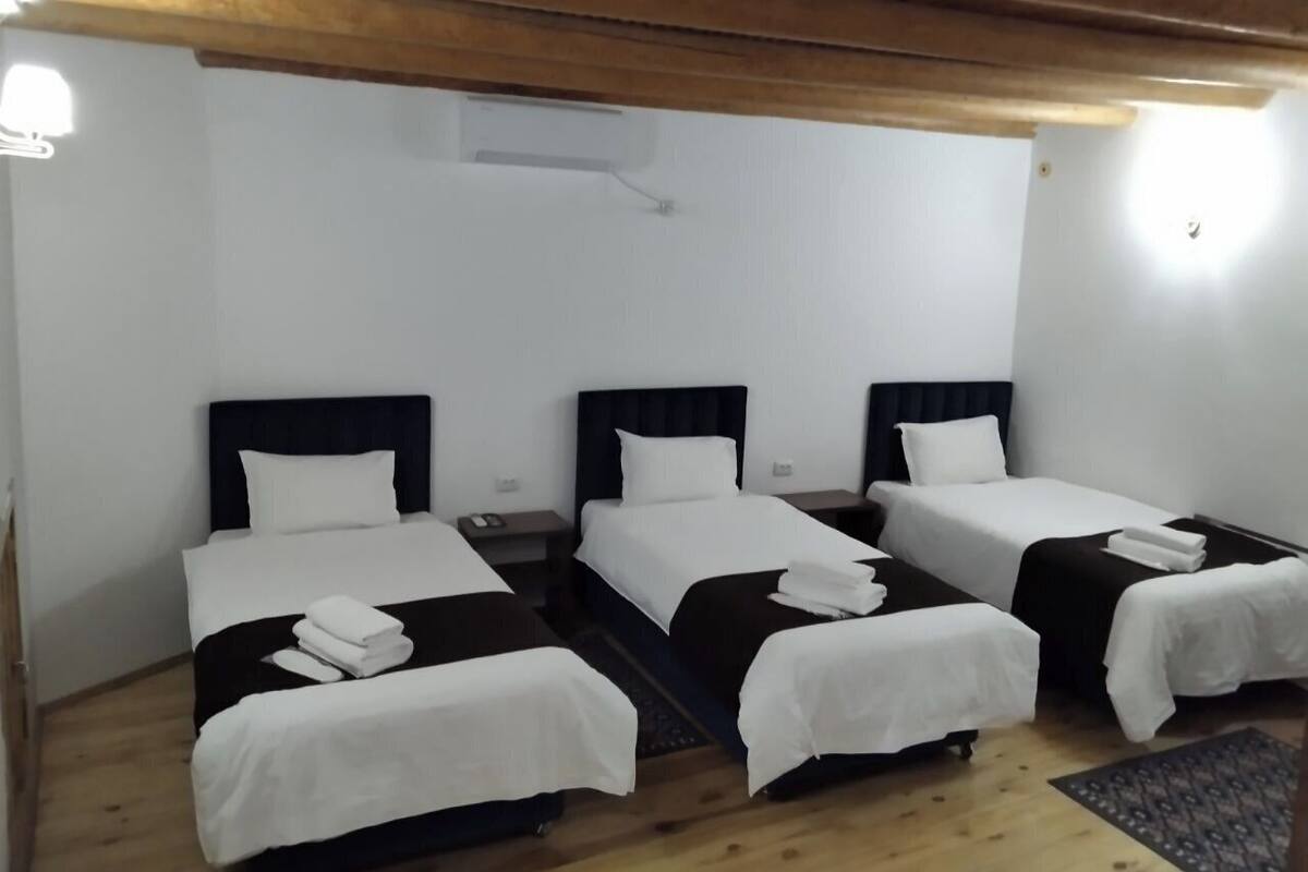 A hotel room with three single beds, each with a white bedspread and a dark headboard. There is a wooden ceiling and a small rug on the floor.