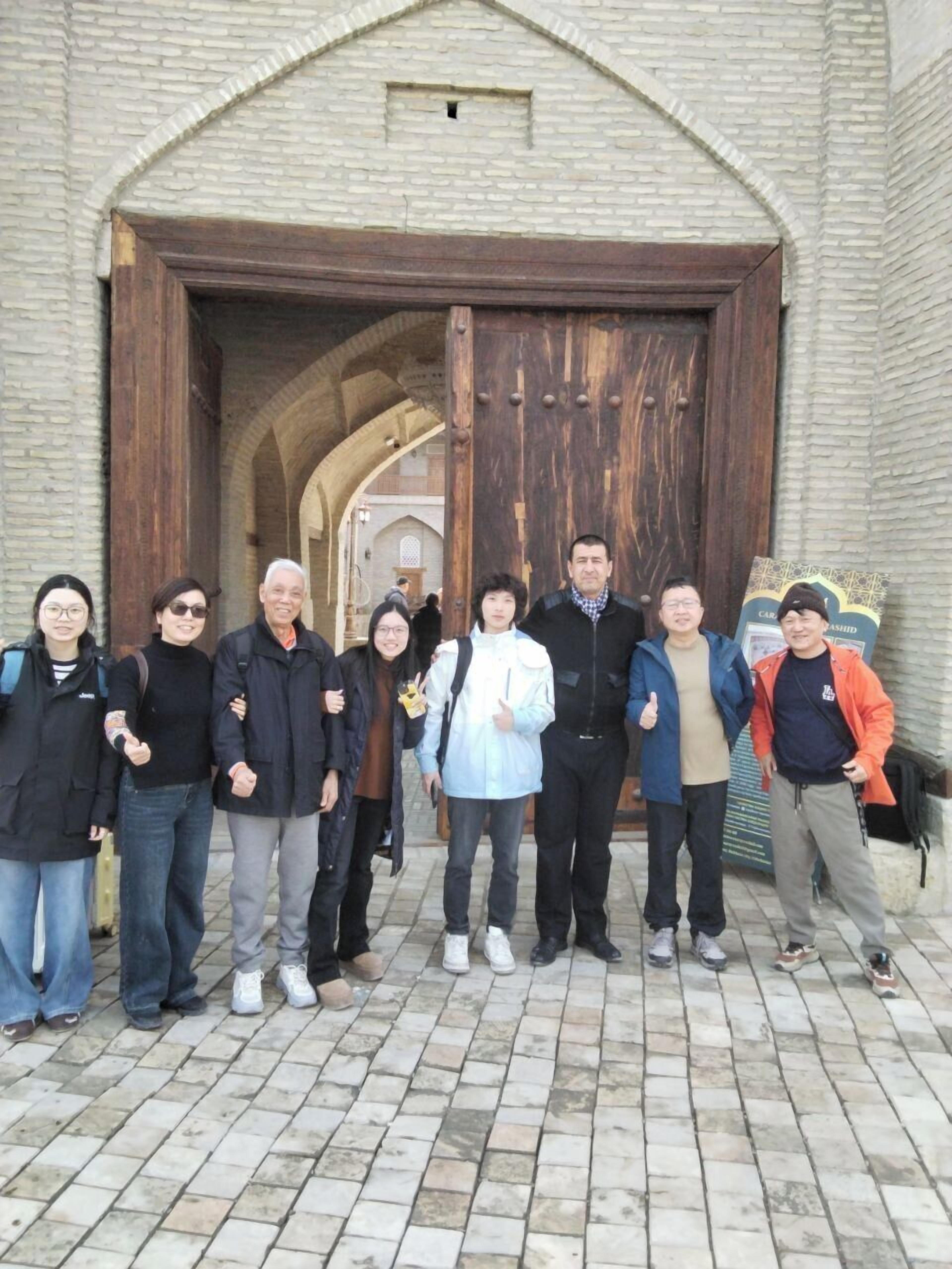 Un grupo de personas de pie frente a una gran puerta de madera con entrada arqueada, sobre un muro de ladrillo.
