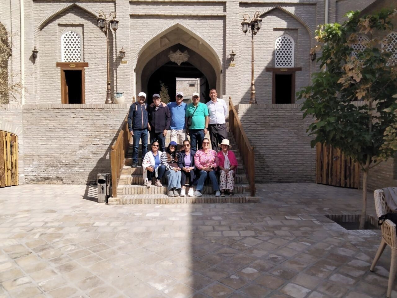 A group of people standing and sitting on stone steps in front of a building with arched windows and a wooden door.