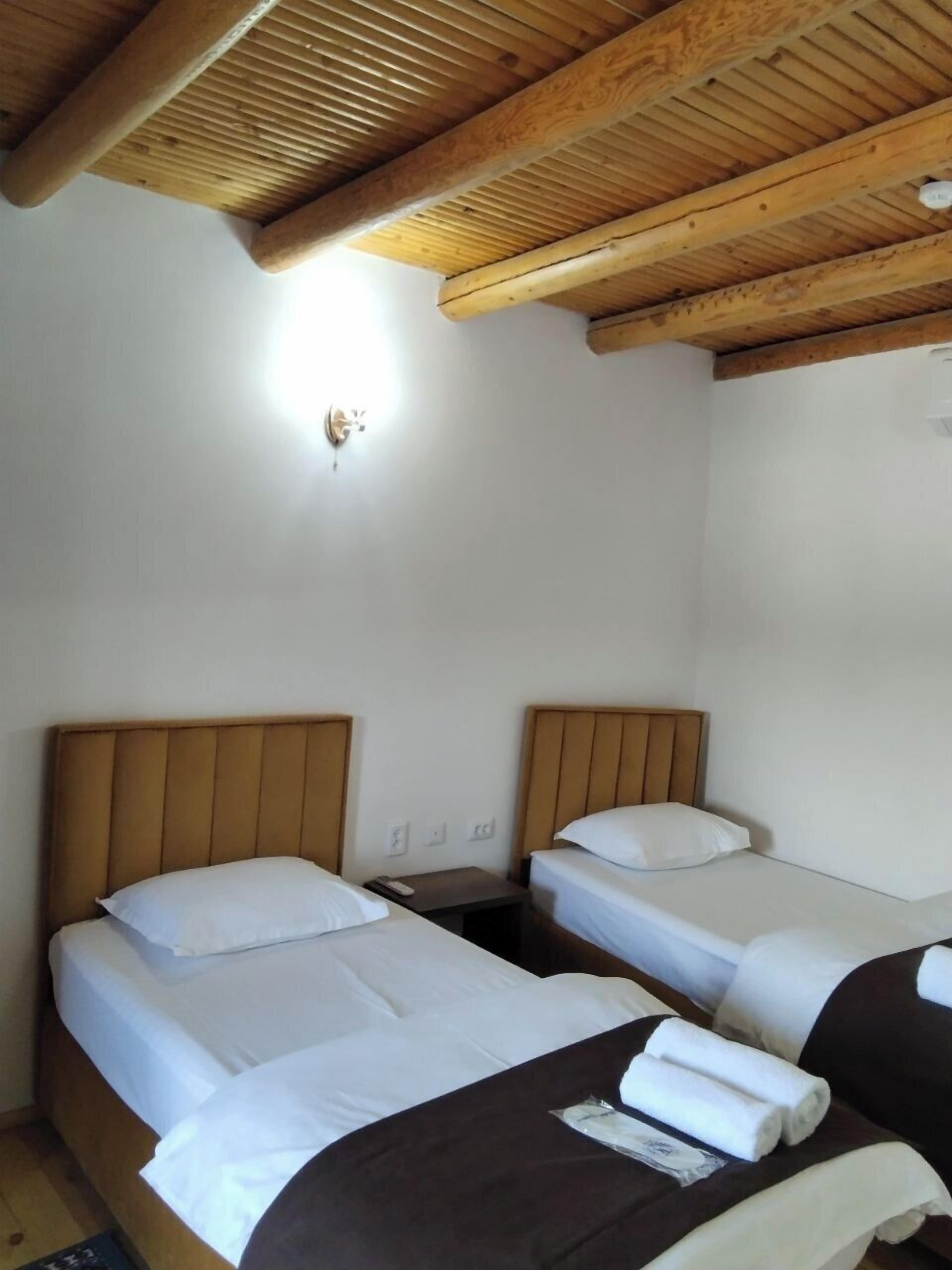 A room with two beds, wooden headboards, white bedding, a small bedside table with a remote control, and a wall-mounted light fixture.