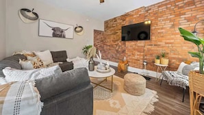 Un salon moderne avec un mur en briques, un ventilateur de plafond, un téléviseur à écran plat et un coin salon confortable avec un canapé et un pouf.