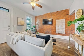 A living room with a brick wall, a ceiling fan, a flat-screen TV, and a large plant.