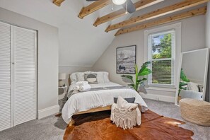 Een slaapkamer met een schuine kap, een bed met witte beddengoed, een groot raam met zonwering, en een plant.