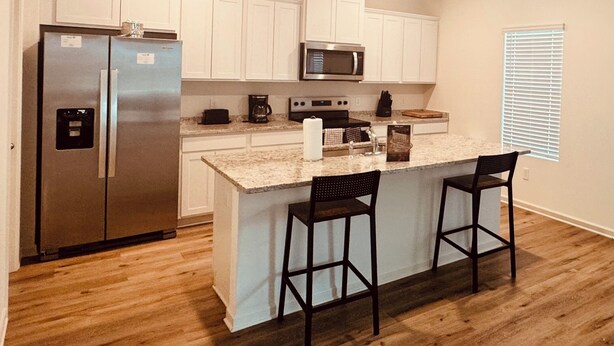 A kitchen with white cabinets, a stainless steel refrigerator, a microwave, a coffee maker, and a small island with two bar stools. - 4-bedroom house in charming Timberline Golf neighborhood with pool (Calera)