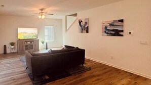 A living room with a dark-colored sofa, wooden flooring, a ceiling fan, and two abstract paintings on the wall. - 4-bedroom house in charming Timberline Golf neighborhood with pool (Calera)