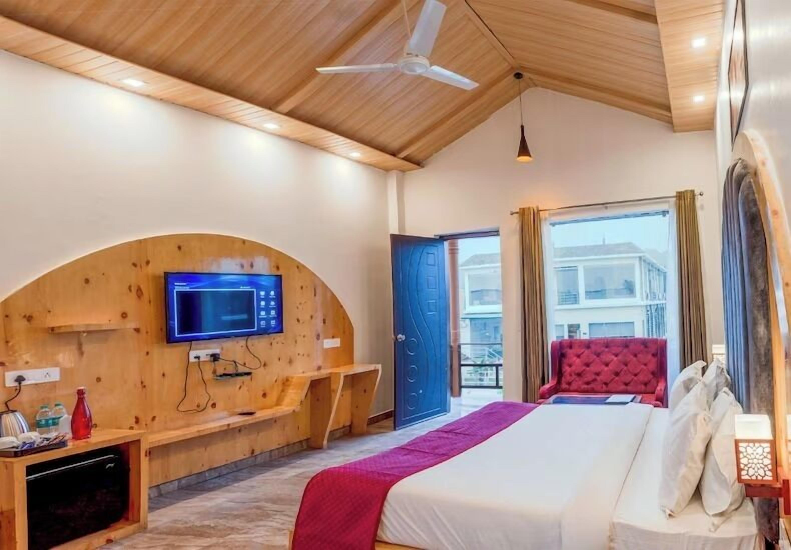 Een moderne hotelkamer met een houten plafond, een flatscreen televisie bevestigd aan een gebogen houten paneel, een bed met een rode hoofdbord en een balkon met uitzicht op gebouwen.