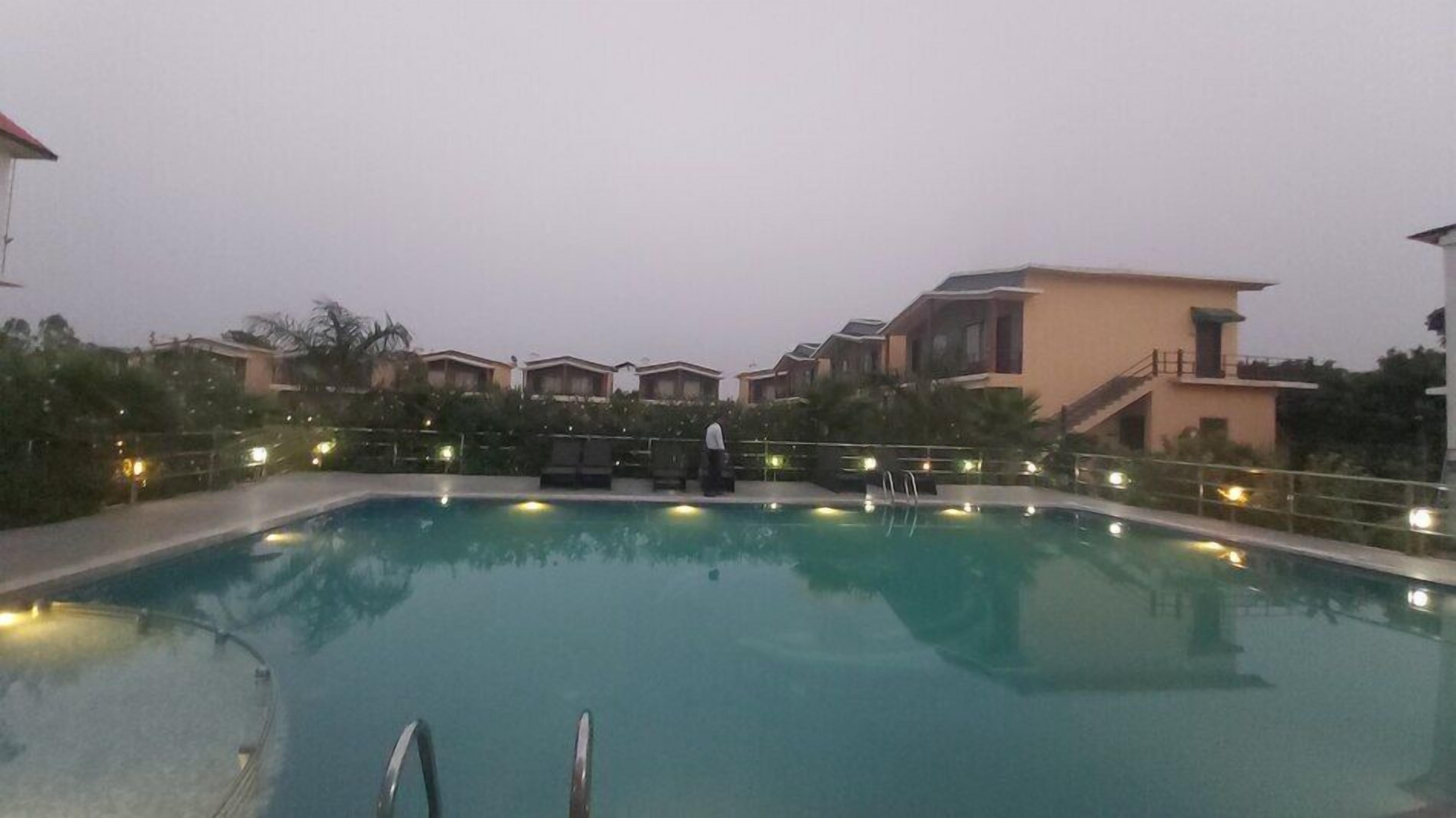 Una piscina con una persona de pie junto al borde, rodeada de edificios y follaje.