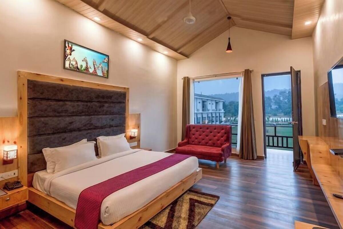 Ein Schlafzimmer mit einem großen Bett, einem roten Sessel, einem Holztisch und Blick auf ein Gebäude durch ein Fenster.