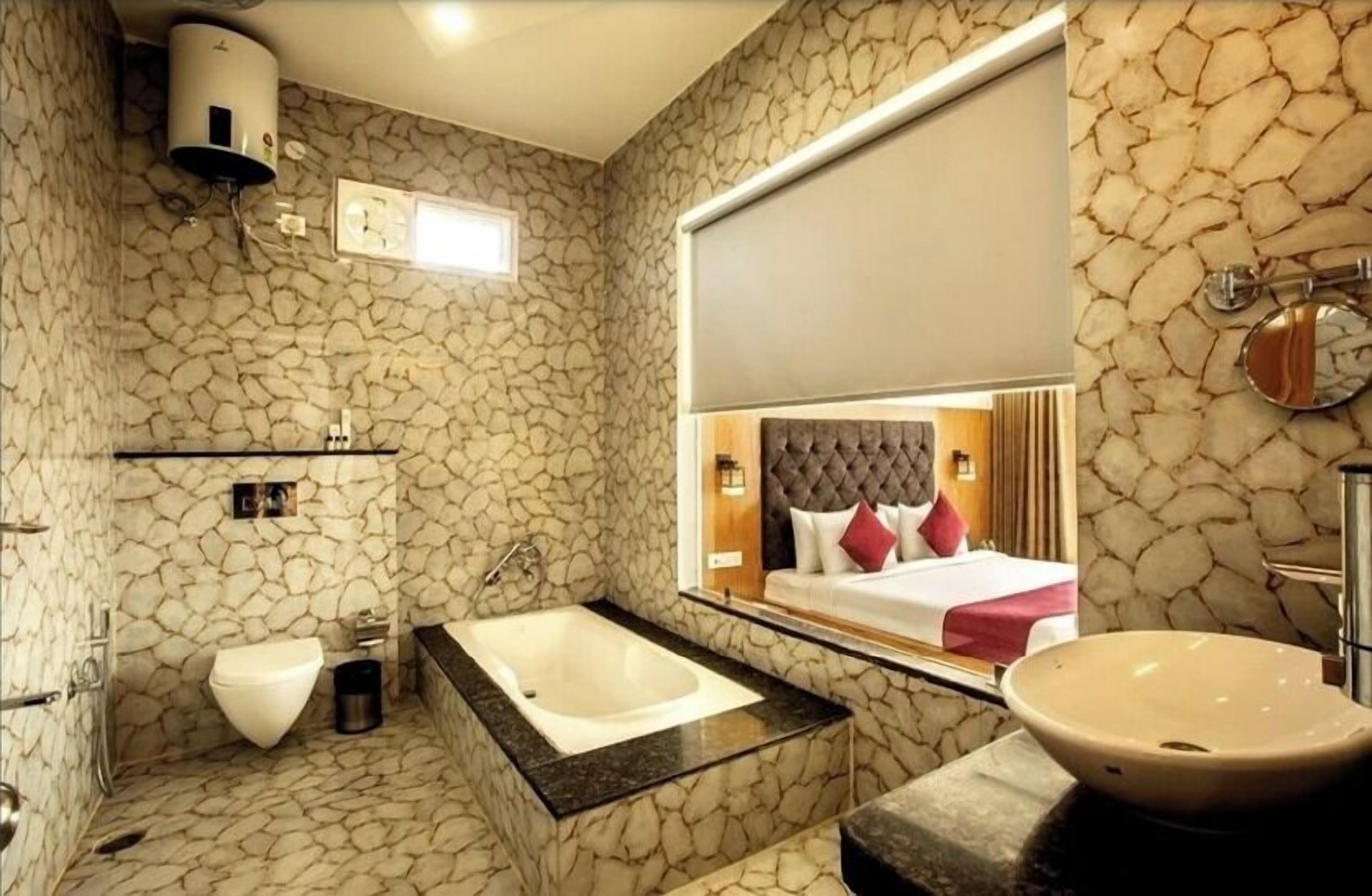 A bathroom with a stone wall, a bathtub, a toilet, and a large mirror reflecting a bed.