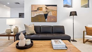 A modern living room with a black leather sofa, a coffee table with decorative items, a floor lamp, and a large abstract painting on the wall. - Spacious 3-Bed Home with Patio Near Beach (Alexandra Headland)