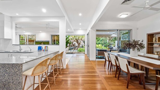 A modern kitchen with a large island, bar stools, and a dining area with wooden chairs. - Spacious 3-Bed Home with Patio Near Beach (Alexandra Headland)