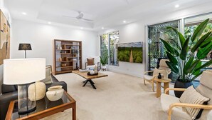 A modern living room with a sofa, coffee table, bookshelf, and large windows offering a view of greenery. - Spacious 3-Bed Home with Patio Near Beach (Alexandra Headland)