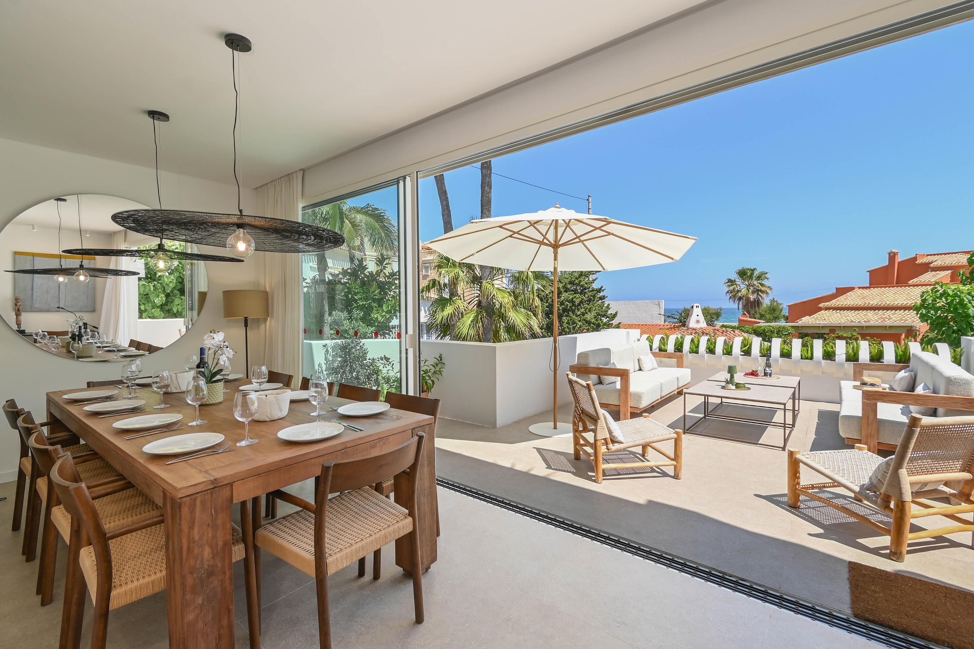 Een eethoek met een houten tafel voor zes personen, omringd door rieten stoelen, en een grote parasol die schaduw biedt.