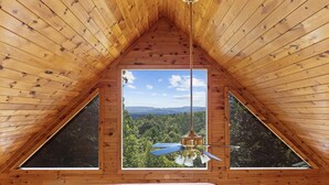 A wooden interior with a triangular ceiling, a ceiling fan, and a window offering a view of a forested landscape.